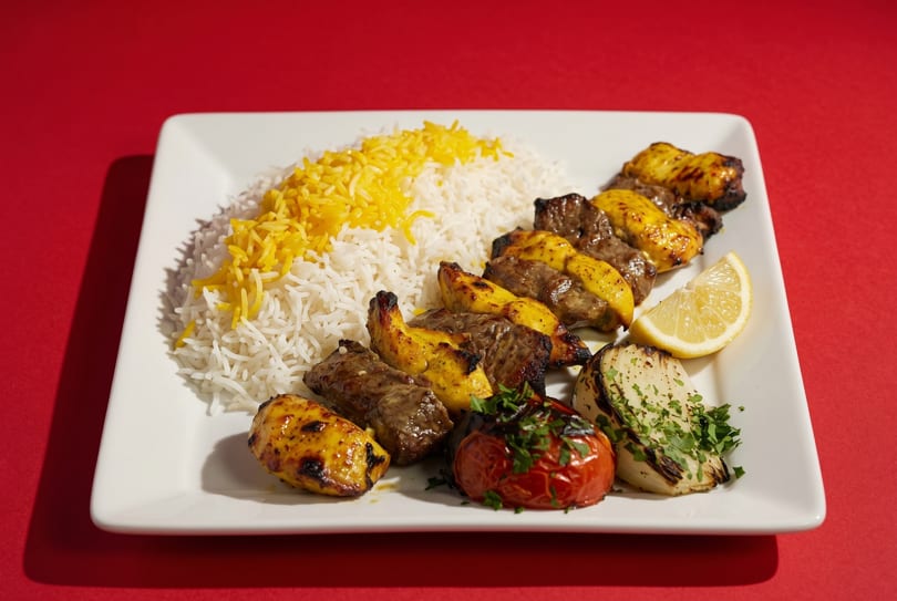A white plate with rice, chicken and beef kabobs, grilled tomato, onion, and a lemon wedge.